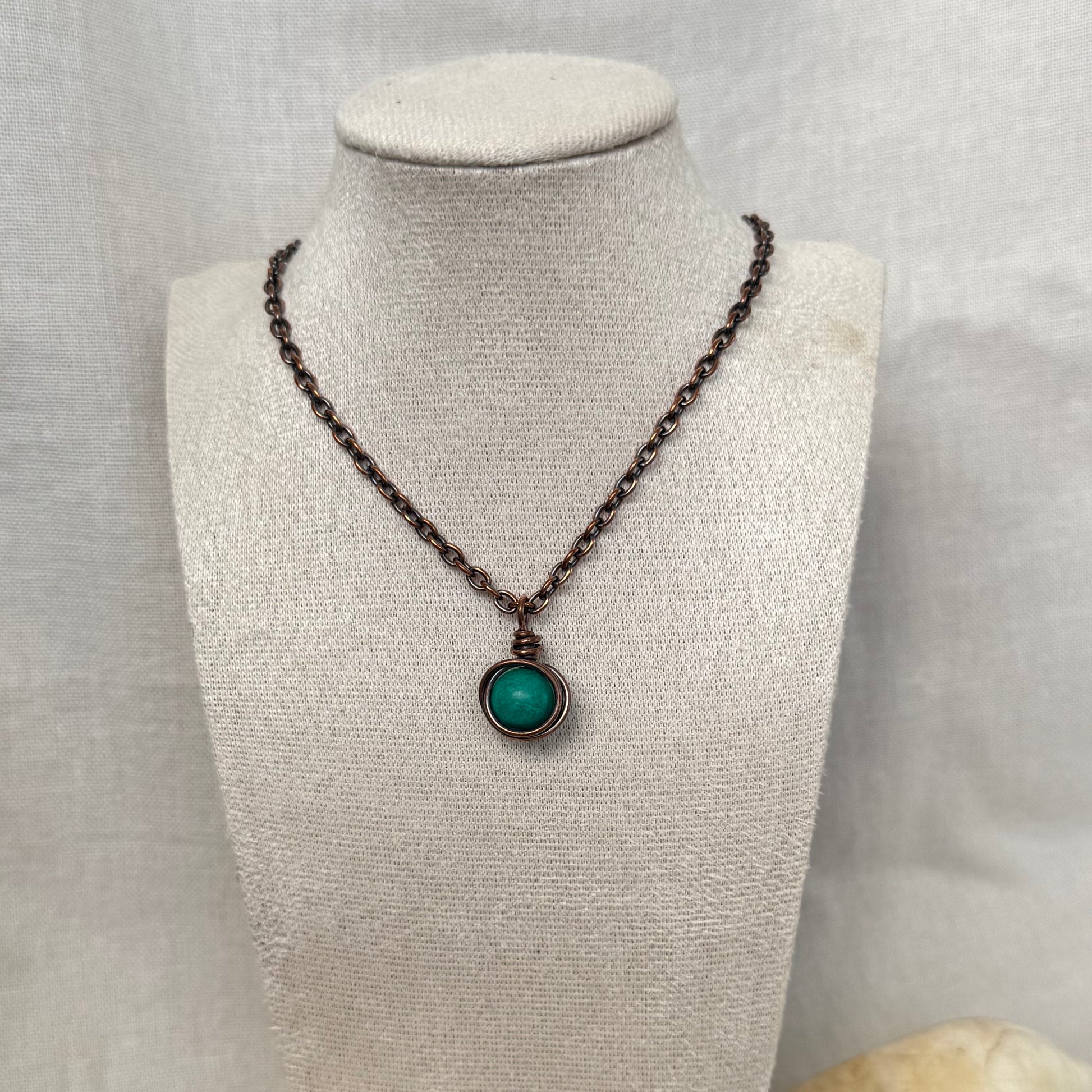 A copper necklace with a copper chain and a round pendant featuring a turquoise round gemstone, displayed on a textured beige fabric jewellery bust.