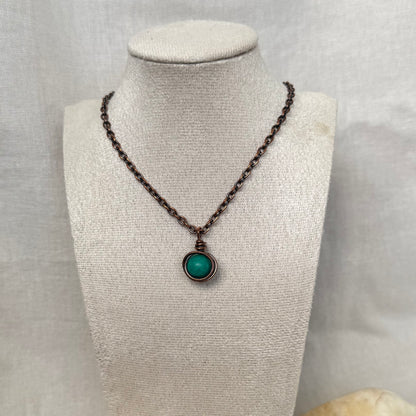 A copper necklace with a copper chain and a round pendant featuring a turquoise round gemstone, displayed on a textured beige fabric jewellery bust.
