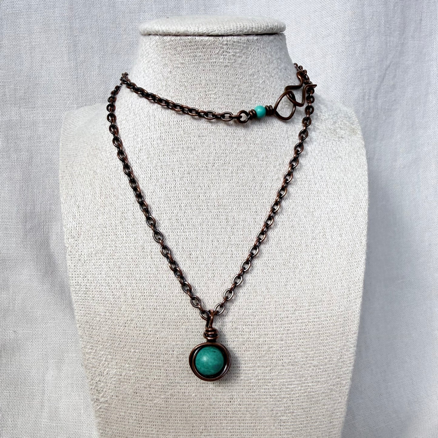 A dark copper chain necklace with a round turquoise pendant, displayed on a textured cream fabric mannequin bust.