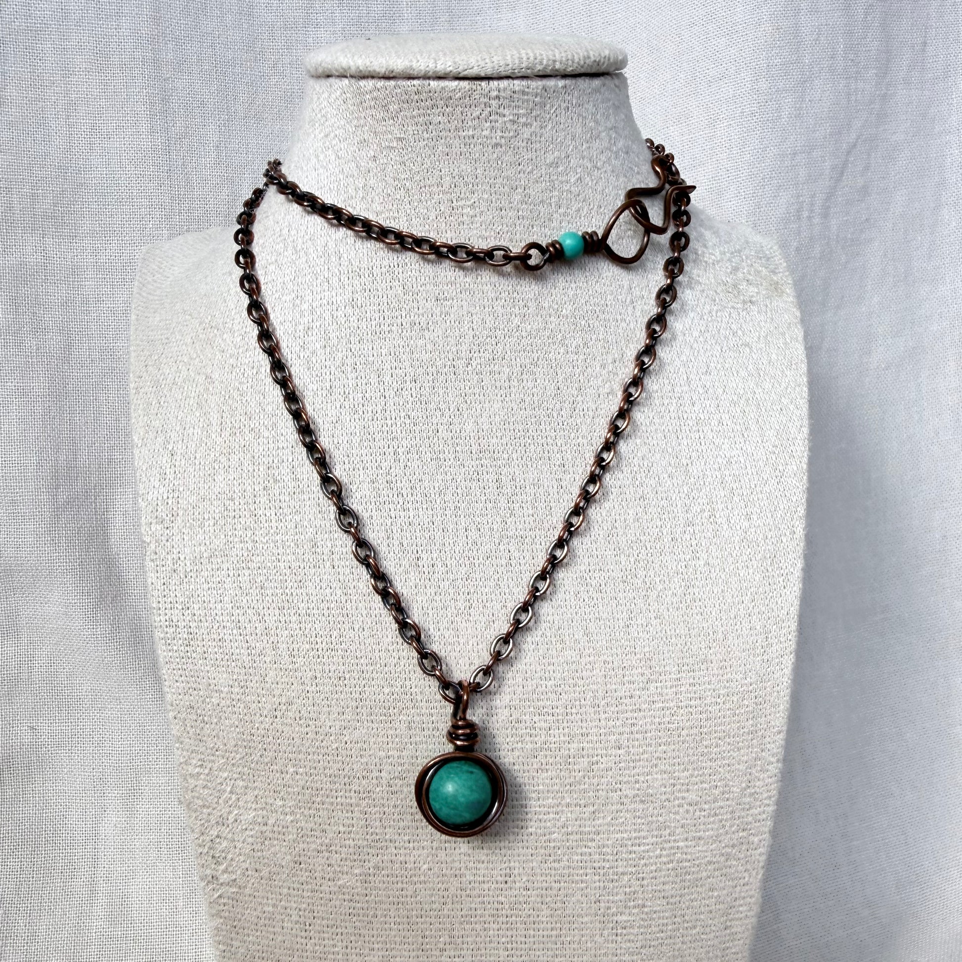 A dark copper chain necklace with a round turquoise pendant, displayed on a textured cream fabric mannequin bust.