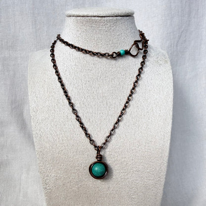 A dark copper chain necklace with a round turquoise pendant, displayed on a textured cream fabric mannequin bust.