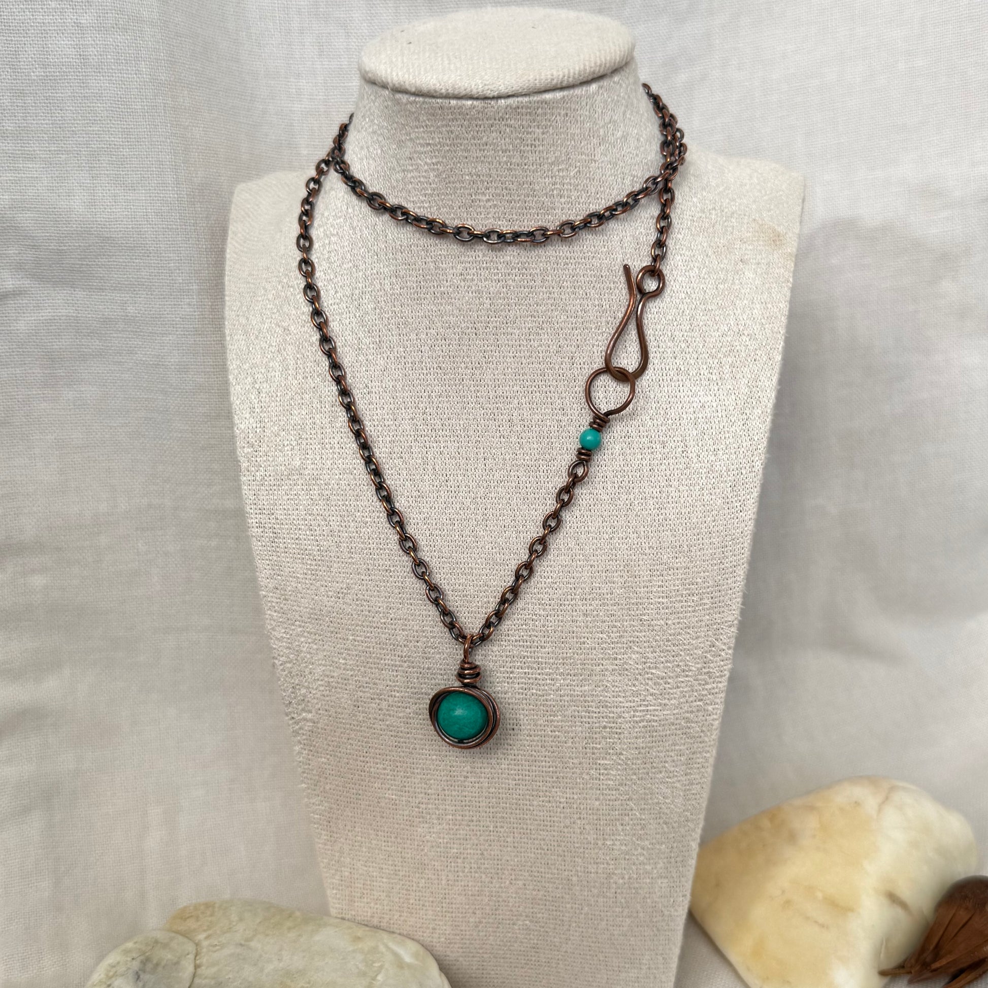 A dark copper chain necklace with a round turquoise bead wrapped in copper wire, displayed on a cream fabric mannequin bust. 