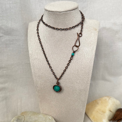A dark copper chain necklace with a round turquoise bead wrapped in copper wire, displayed on a cream fabric mannequin bust. 