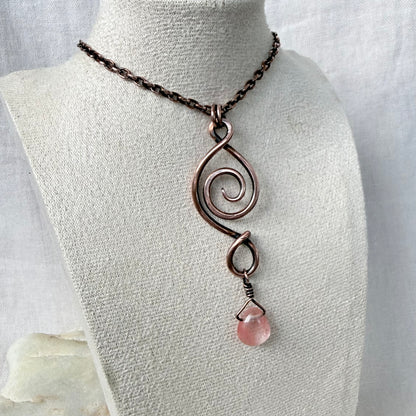 A handmade copper spiral pendant necklace with a dangling pink cherry quartz stone, displayed on a textured off-white fabric mannequin bust.