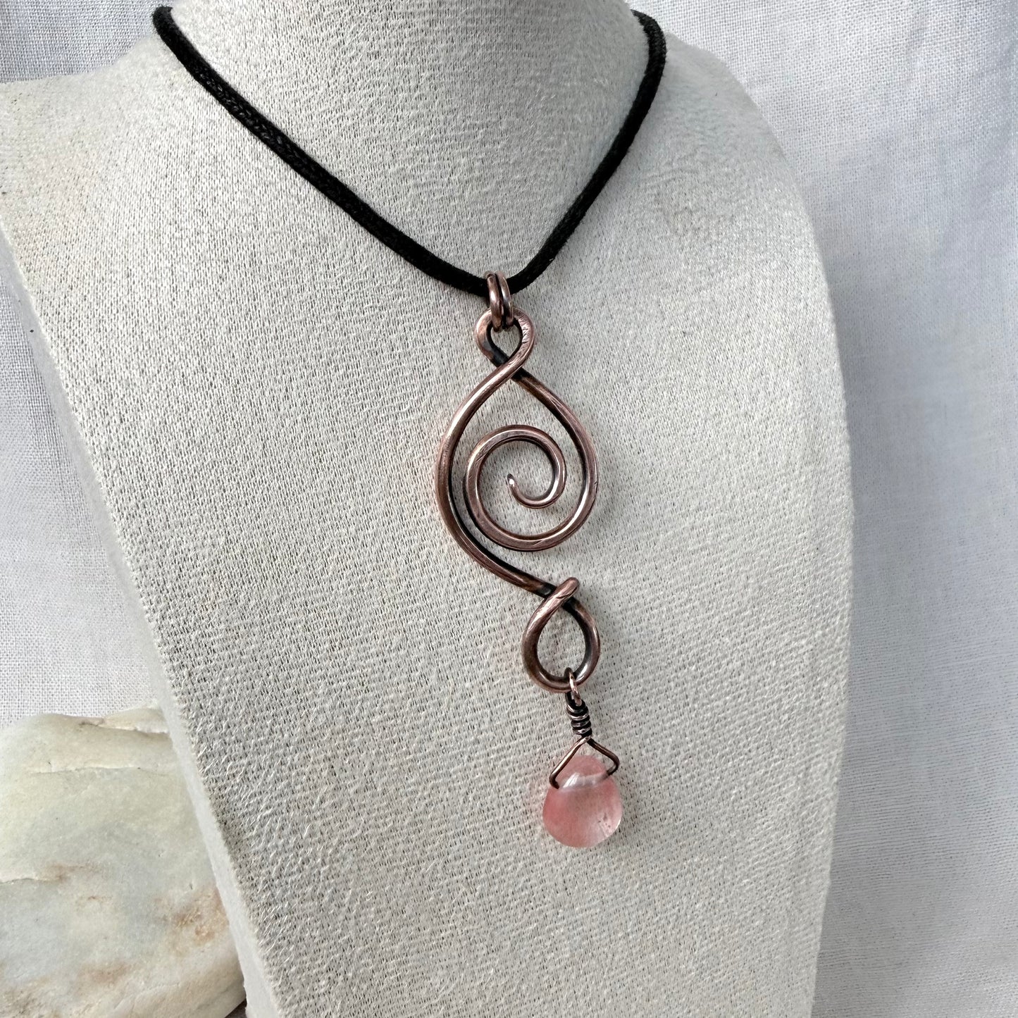 A handmade wire-wrapped pendant necklace with a spiral design and a dangling cherry quartz stone, displayed on a textured white mannequin bust.
