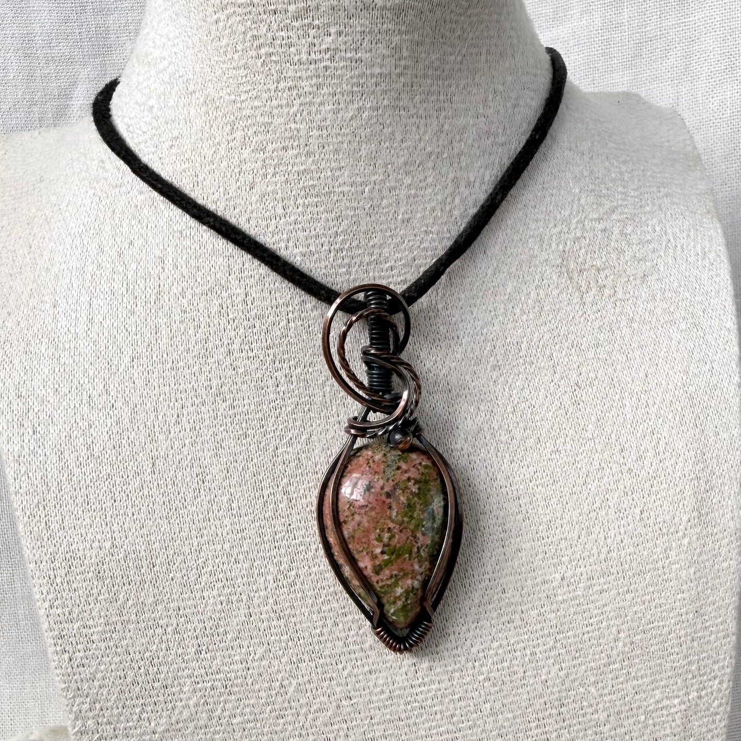 A teardrop-shaped pendant necklace with a polished pink and green Unakite stone wrapped in copper, displayed on a textured white mannequin bust with a black cord.