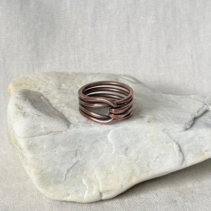 A multi-strand copper wire ring with a twisted center detail, displayed on a light gray stone against a neutral linen background.
