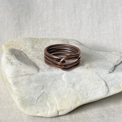 A handmade multi-band ring made of twisted copper-colored metal wires, resting on a light gray stone slab against a neutral linen background.