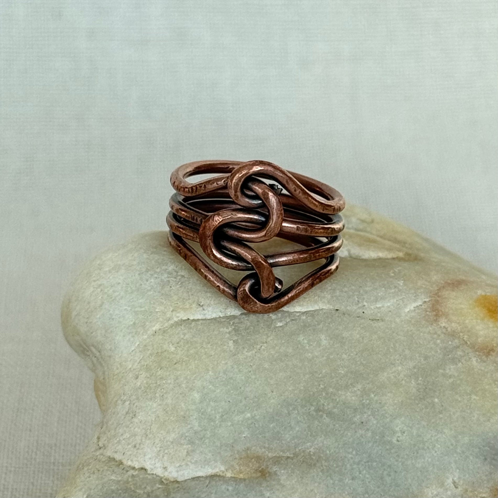A handmade copper wire ring with a knotted, interwoven design, displayed on a smooth light-colored stone against a neutral textured background.