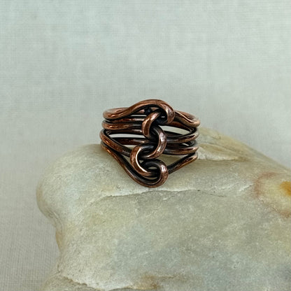 A handmade copper ring with a knotted, interwoven design, resting on a smooth light-colored stone against a neutral fabric background.
