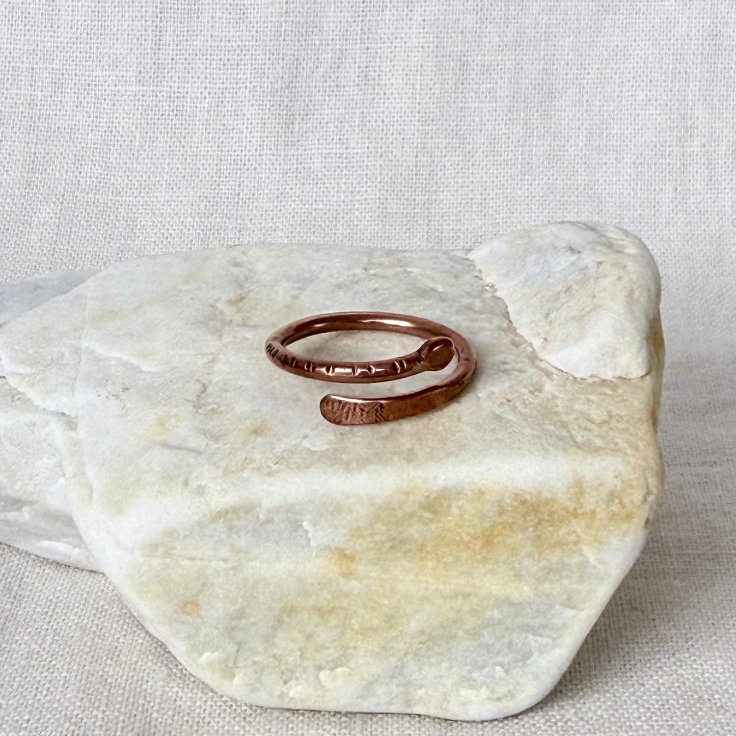 A copper-toned open wrap ring with hammered texture details, displayed on a light beige stone against a neutral linen background.