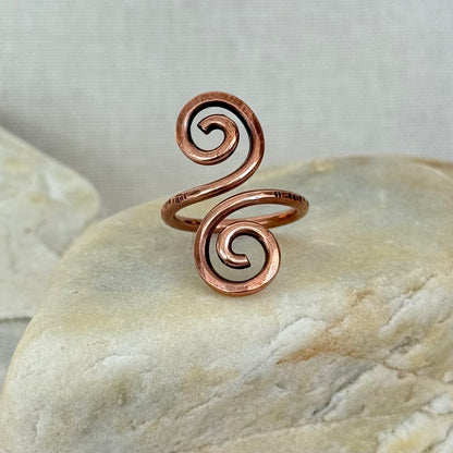 A copper spiral wrap ring displayed on a smooth beige stone against a neutral textured background.