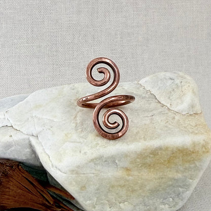 A copper spiral ring with two curled ends, displayed on a textured white stone against a neutral beige fabric background.
