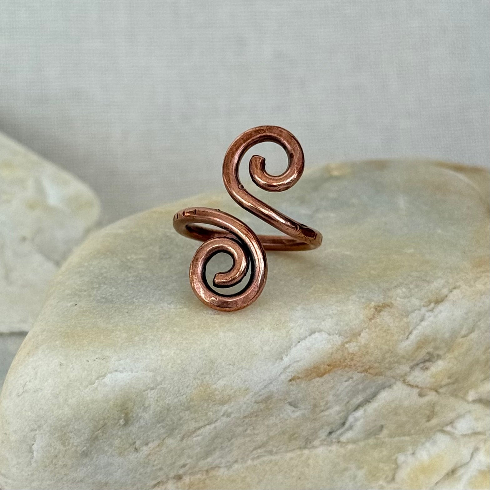 A copper spiral wrap ring displayed on a smooth beige stone against a neutral light gray background.
