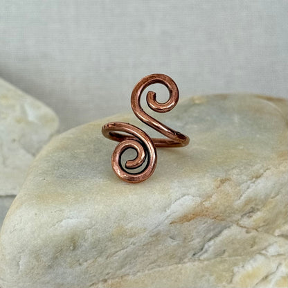 A copper spiral wrap ring displayed on a smooth beige stone against a neutral light gray background.