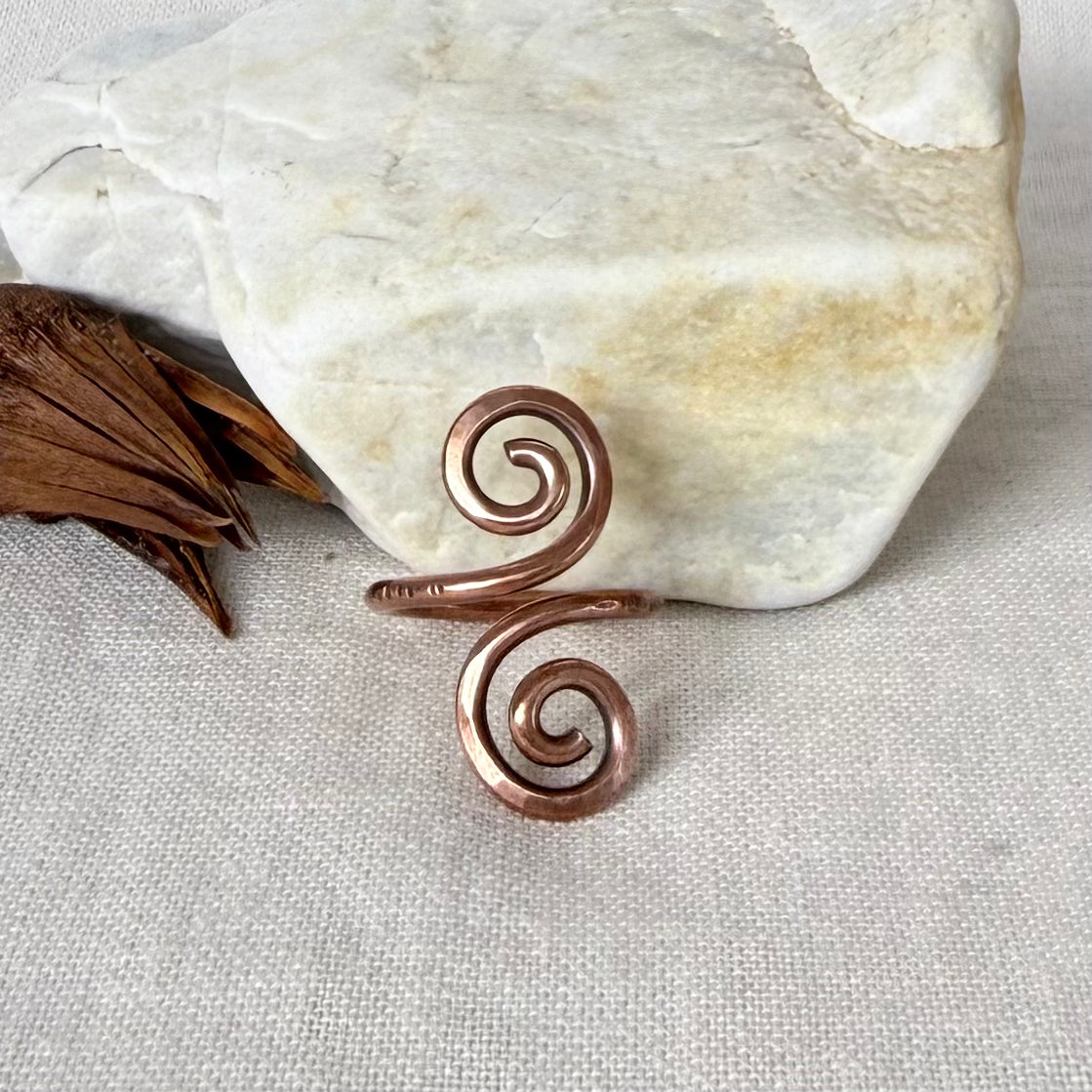 A copper spiral ring with a hammered texture, styled on a beige linen fabric next to a white stone and dried brown leaves.