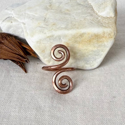A copper spiral ring with a hammered texture, styled on a beige linen fabric next to a white stone and dried brown leaves.