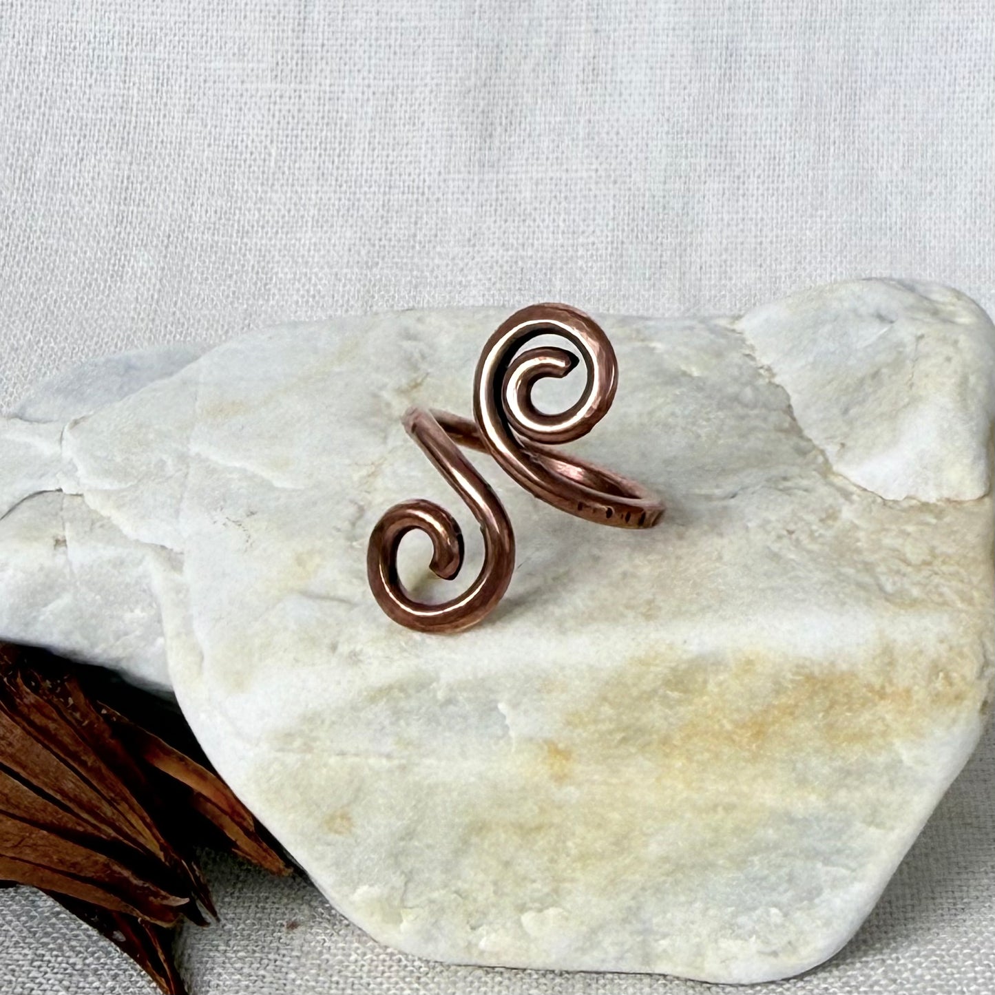 A copper-toned spiral wrap ring displayed on a smooth white stone against a light beige fabric background.