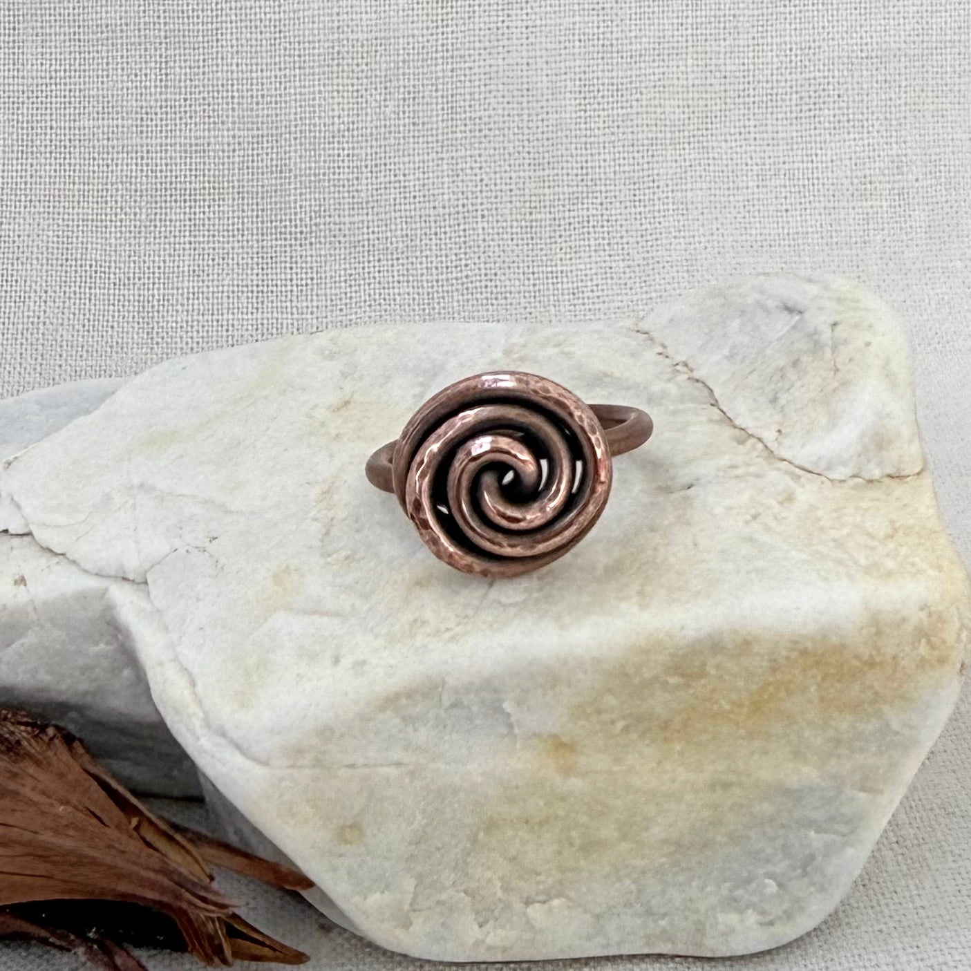 A hammered copper spiral ring displayed on a light-colored stone against a neutral textured fabric background.