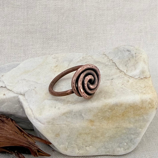 A handmade copper spiral ring resting on a light-colored stone against a neutral linen background.