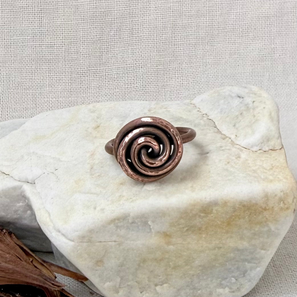 A handmade copper spiral ring displayed on a light-colored stone against a neutral textured fabric background.