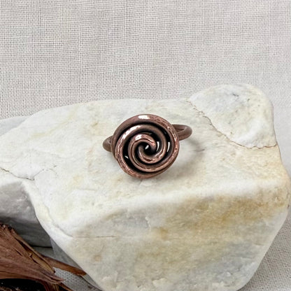 A handmade copper spiral ring displayed on a light-colored stone against a neutral textured fabric background.