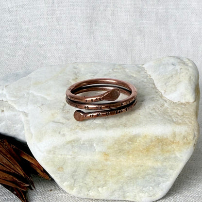 A copper wrap ring with hammer texture details, displayed on a light stone surface against a neutral fabric background.